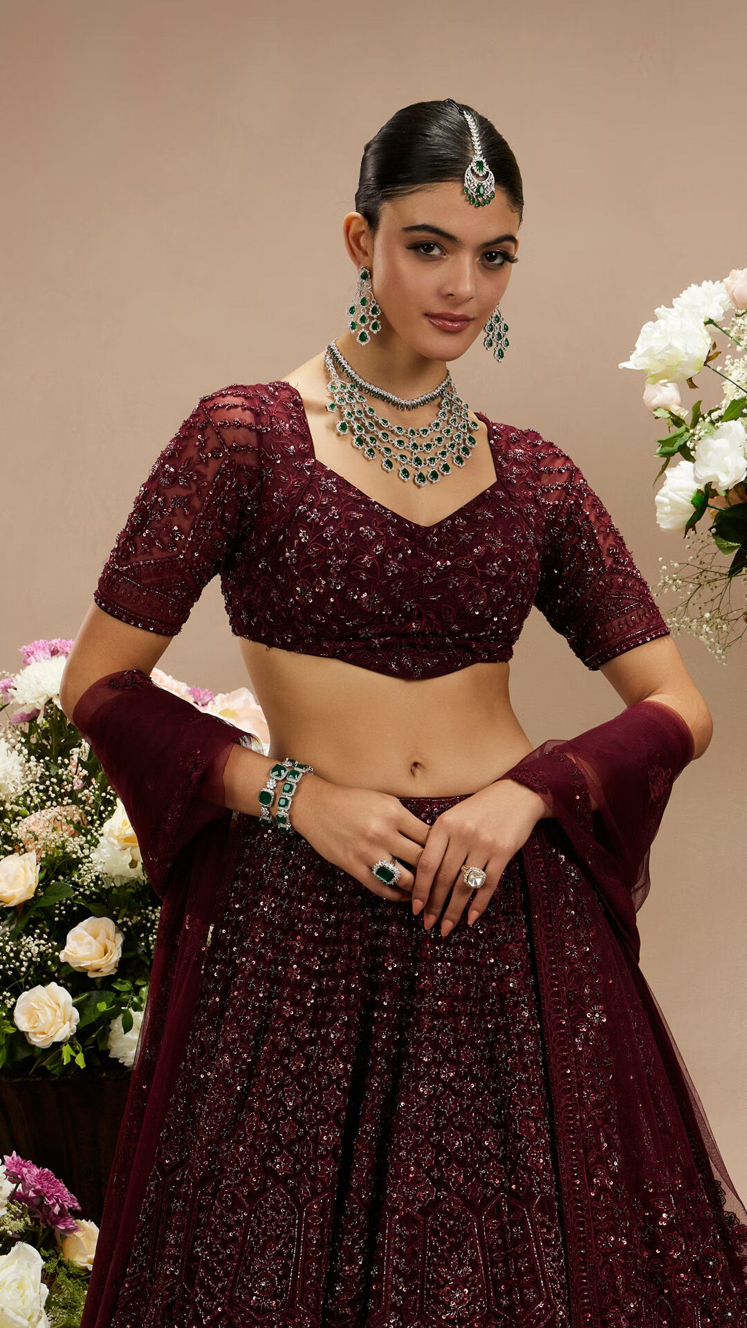 Woman modeling a stunning maroon sequined lehenga and embroidered blouse, complete with emerald jewelry for a regal bridal look.